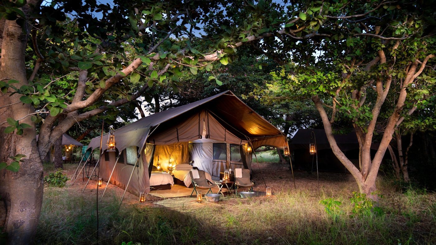 Safari tent exterior lit at dusk among trees at &Beyond Chobe Under Canvas
