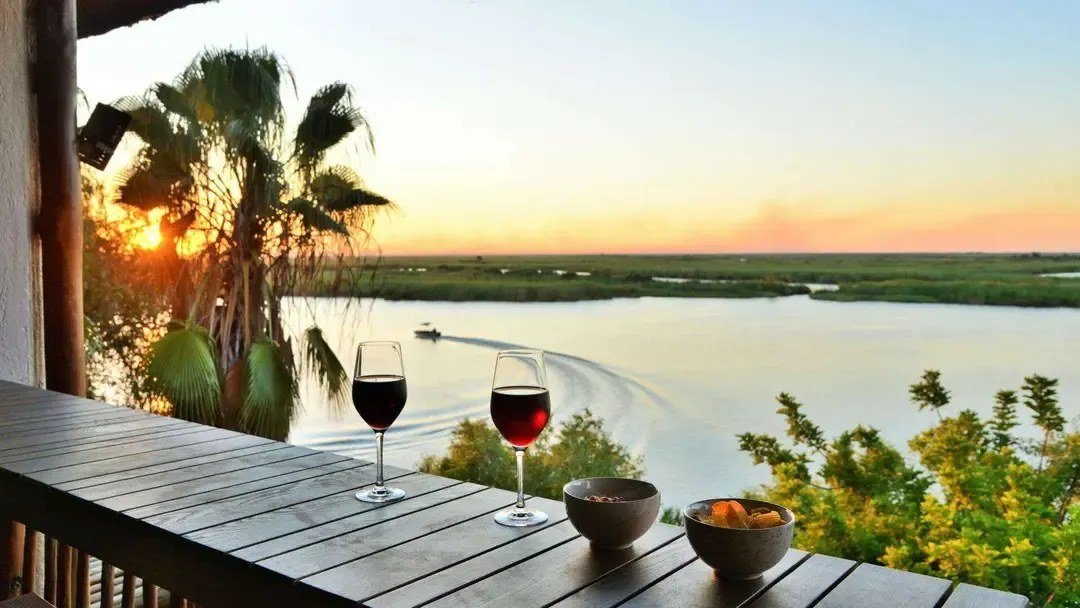 Private balcony with wine glasses overlooking the sunset river at Cresta Mowana Safari Resort & Spa