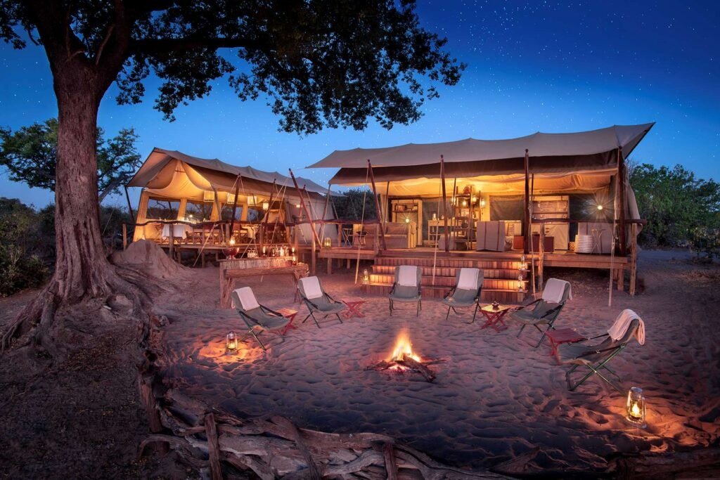 Campfire and lounge chairs under a large tree at dusk at Linyanti Expeditions, Chobe