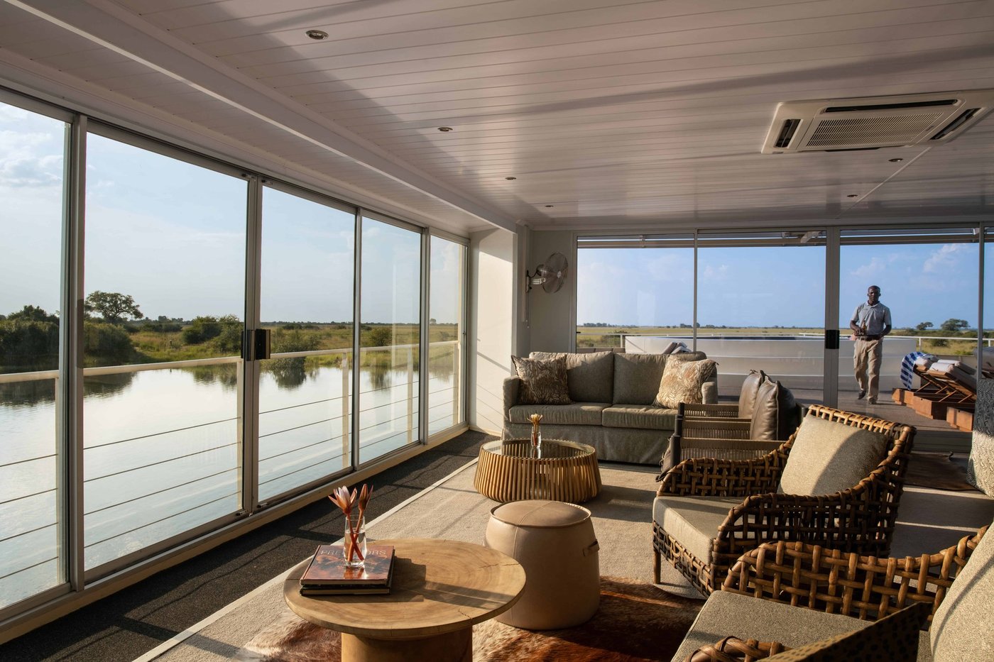 Lounge with wicker furniture and large river-view windows at Zambezi Queen